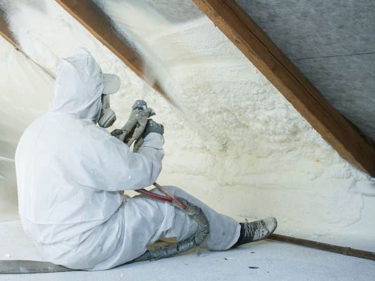 Worker in protective gear applying spray foam insulation in attic space.