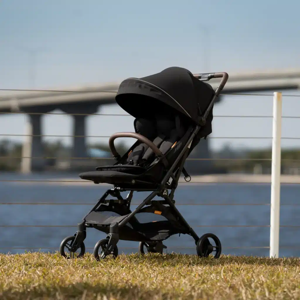 Lightweight black baby stroller on grass by a waterfront and bridge, ideal for outdoor adventures.
