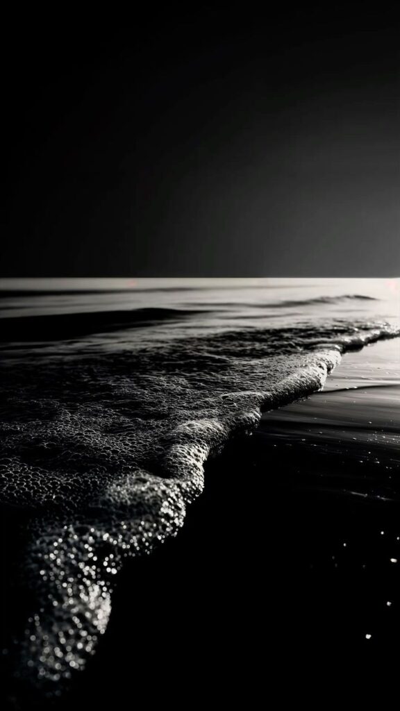 Black and white photo of gentle ocean waves creating foam under a dark sky.