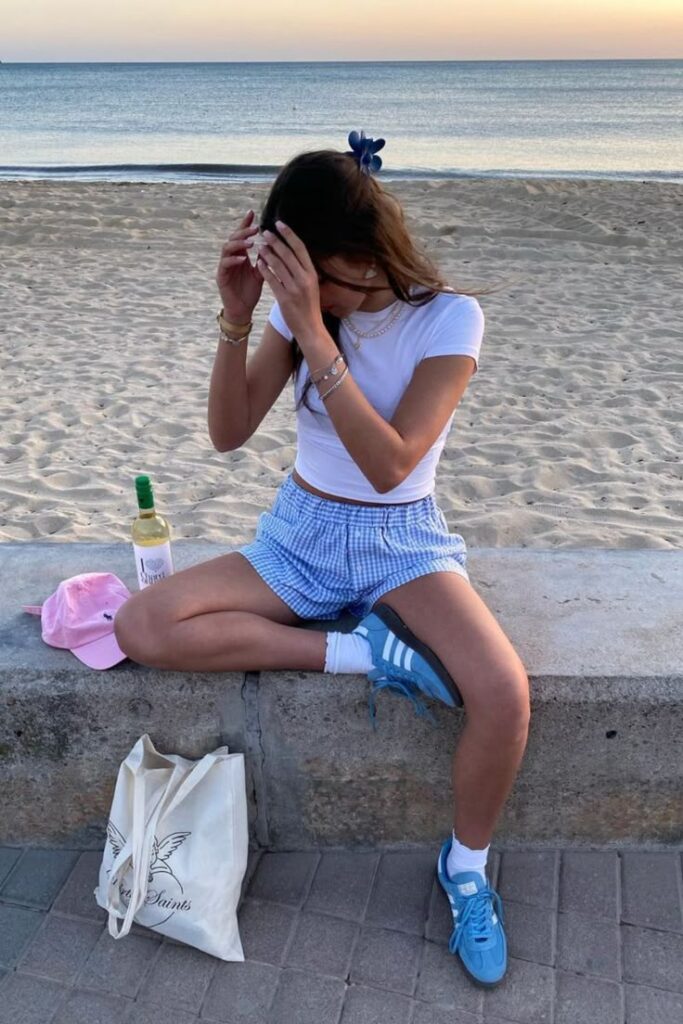 Woman sitting on beach wall at sunset with wine, tote bag, and sneakers, adjusting hair.