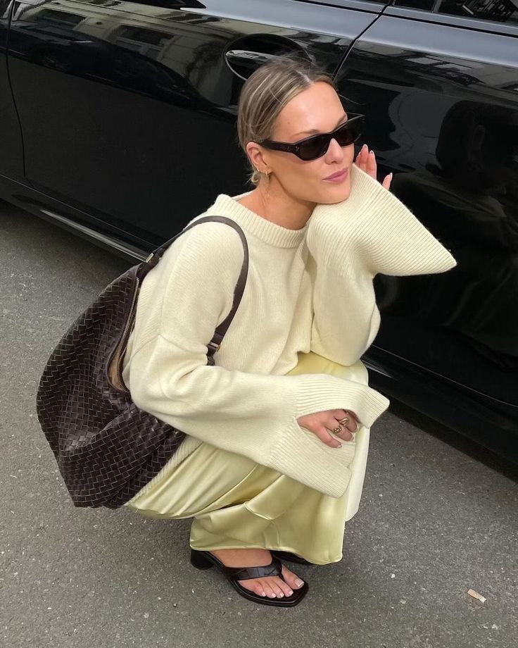 Woman in stylish outfit with sweater, sunglasses, and large bag kneeling by a black car on the street.