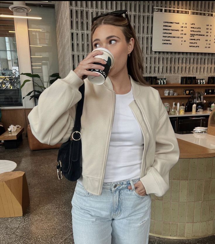 Woman in a café sipping coffee, wearing a beige jacket and jeans, with menu board in the background.