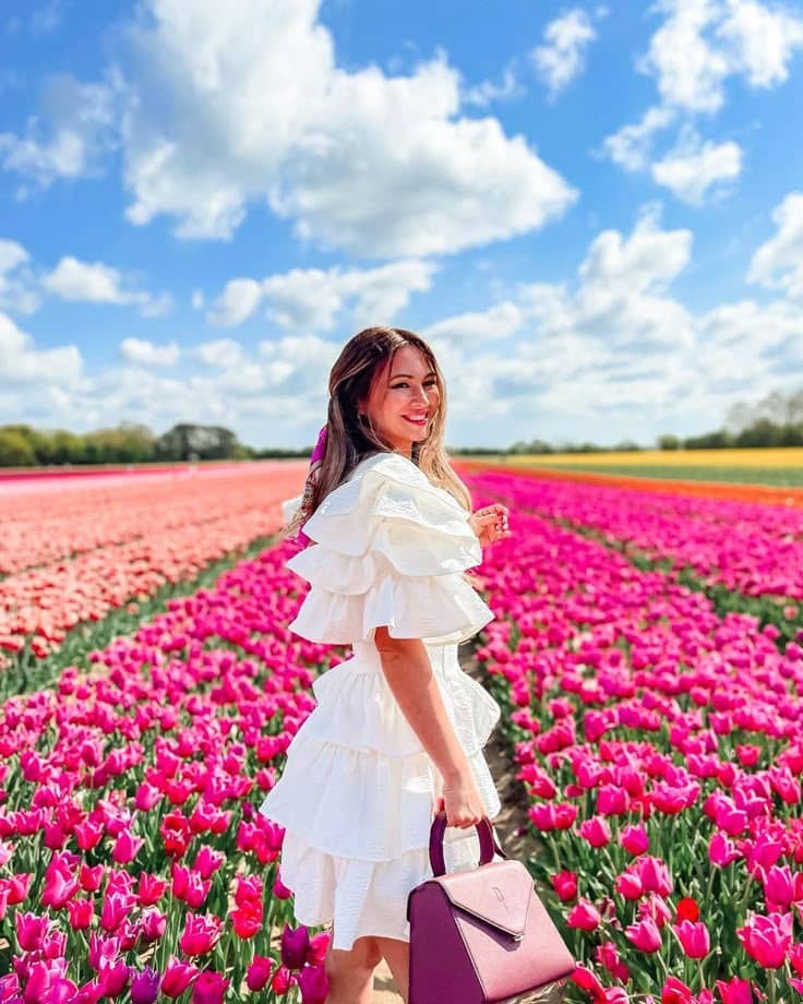 1f6c2ce8-97c9-4d98-8e62-f8f5b9136cdf Woman in white dress smiling amidst vibrant pink tulip fields under a blue sky with fluffy clouds. | Sky Rye Design Woman in white dress smiling amidst vibrant pink tulip fields under a blue sky with fluffy clouds.