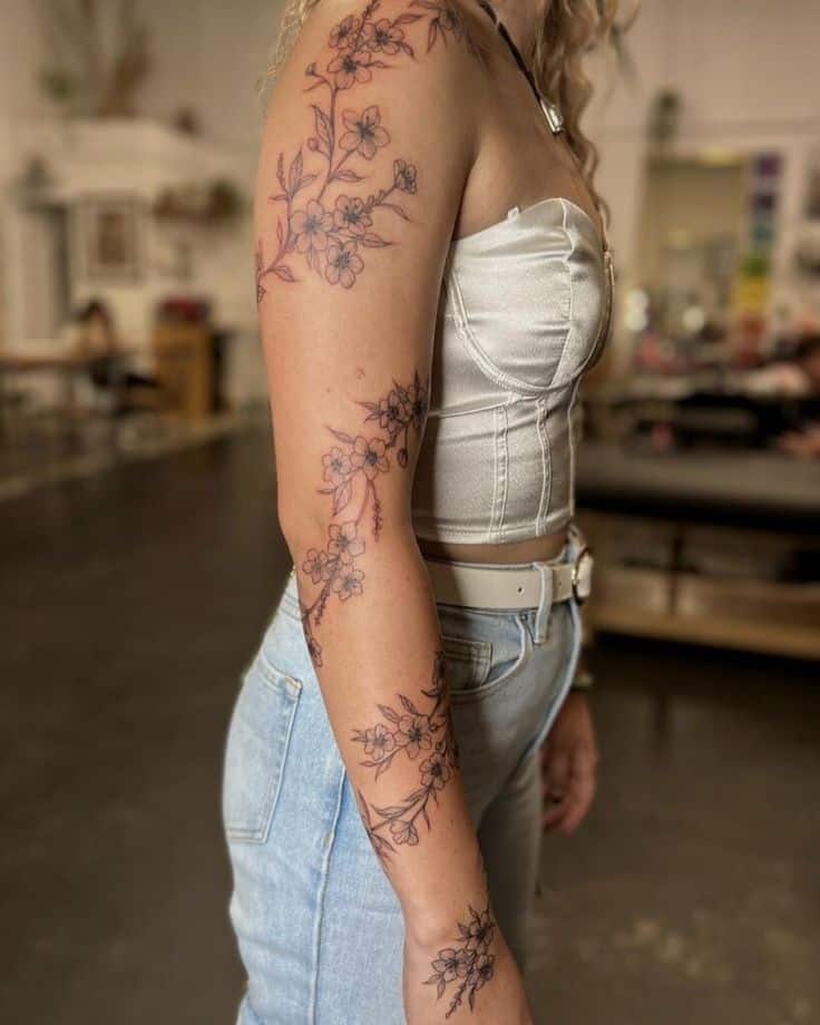 Woman with floral arm tattoos, wearing a white corset top and denim jeans, in a tattoo studio setting.