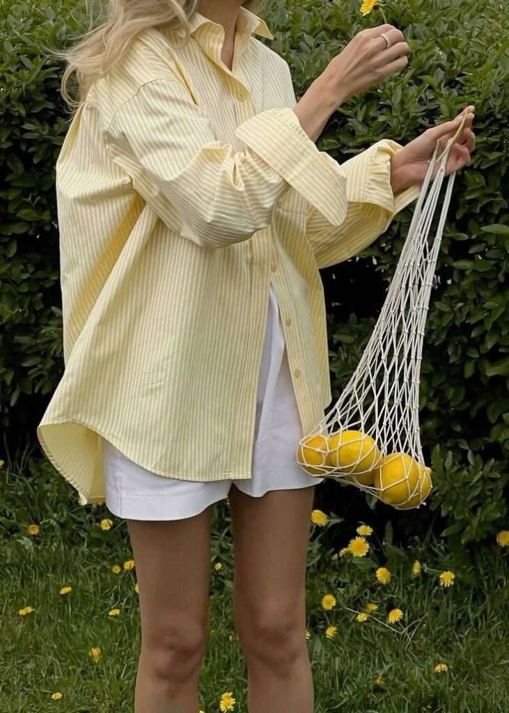 Woman in yellow shirt holding mesh bag with lemons in a garden, surrounded by greenery and flowers.