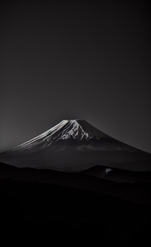 Black and white image of Mount Fuji at night, with snow-capped peak and dark mountainous foreground.
