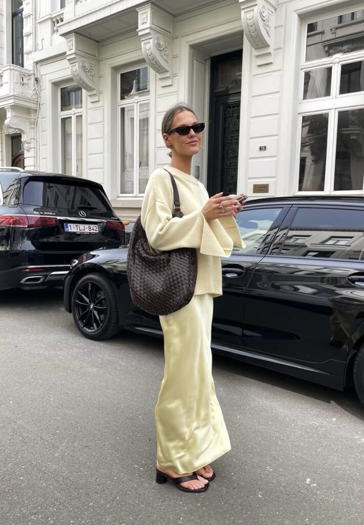 Stylish woman in beige outfit with sunglasses and handbag, standing on street with cars and elegant buildings.