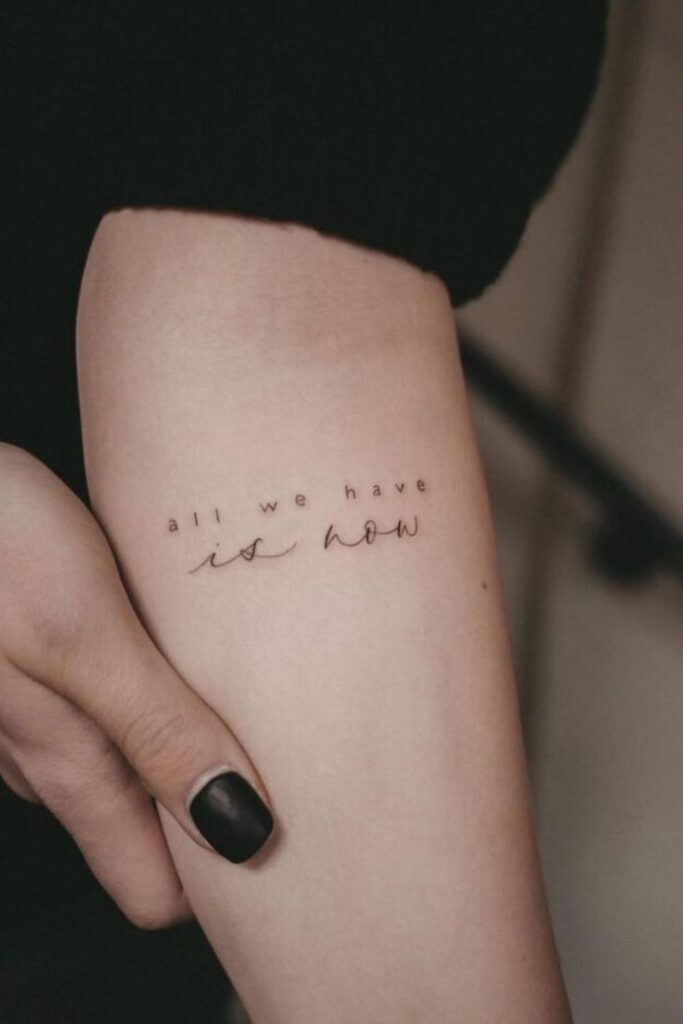 Minimalist tattoo on forearm reading all we have is now, with black painted nails visible beside it.