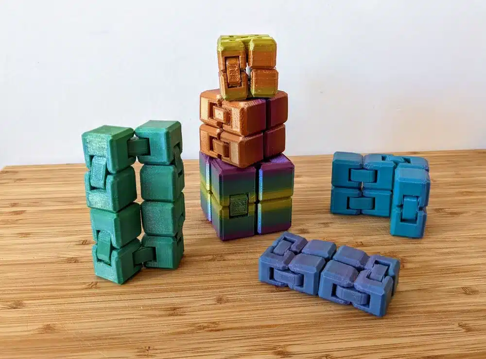 2023-09-25_46f0a05f3505b Colorful 3D-printed fidget cubes on a wooden surface, featuring various stacked and laid out designs. | Sky Rye Design Colorful 3D-printed fidget cubes on a wooden surface, featuring various stacked and laid out designs.