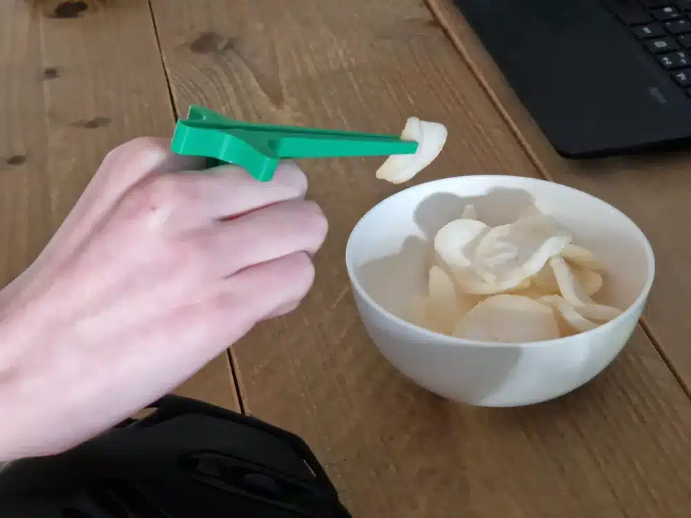 Hand using a green tool to pick up potato chips from white bowl on wooden table near a laptop.