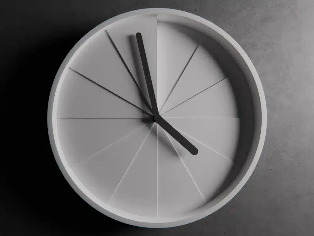 Minimalist clock with black hands and white geometric face against a dark background, viewed from above.