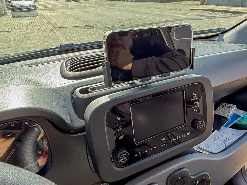 Phone mounted on car dashboard displaying reflection, interior view with steering wheel and controls visible.