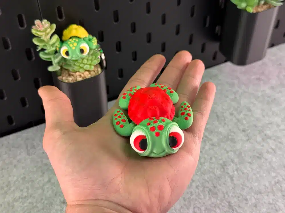 A hand holding a colorful, cartoon-like 3D-printed turtle toy with a succulent plant in the background.