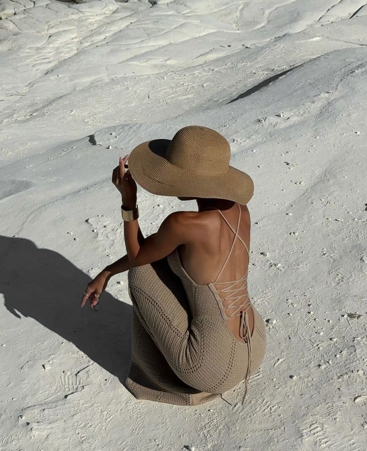 Woman in a backless crochet dress and sun hat sitting on a textured sandy surface, capturing a serene summer moment.