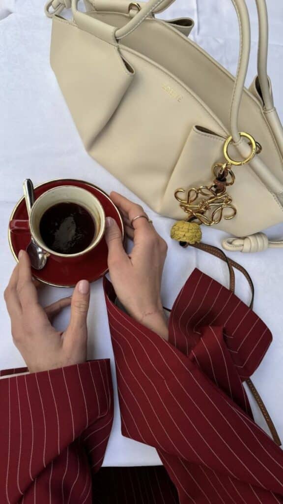 Stylish red-sleeved person holding coffee next to beige designer handbag on a white tablecloth. aesthetic photography 
