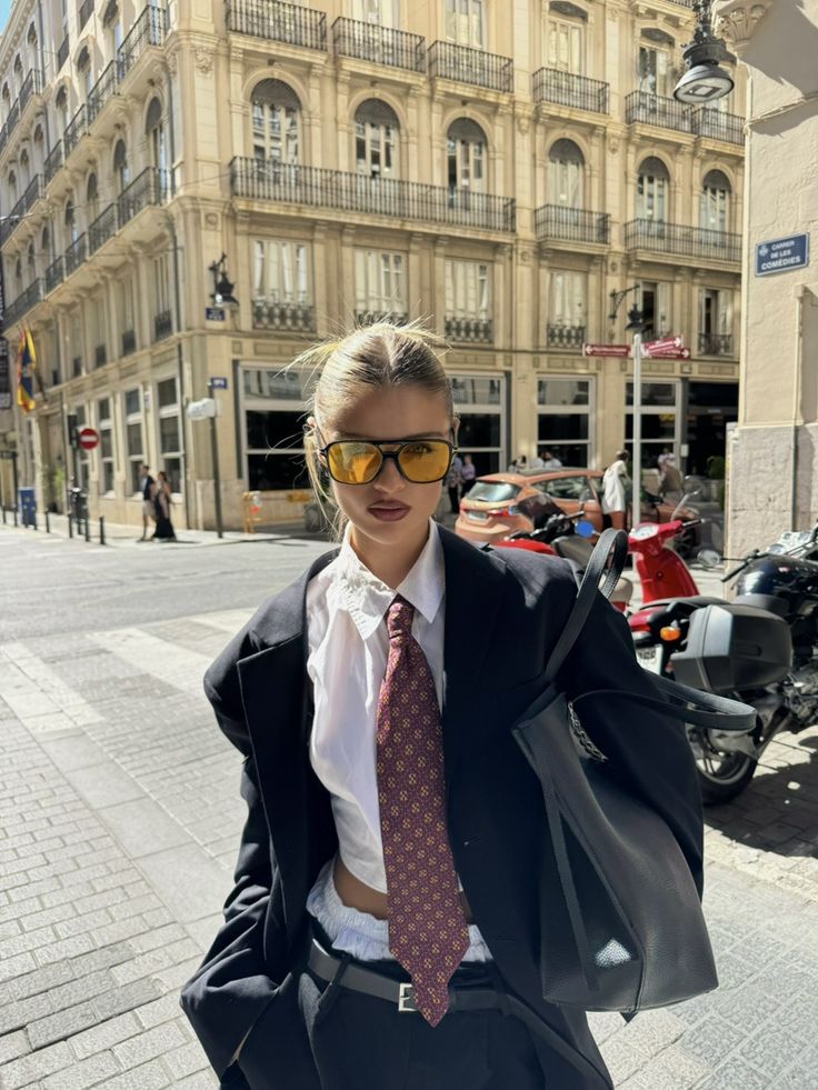 Stylish person in a suit and tie with sunglasses, standing on a city street near a historic building.