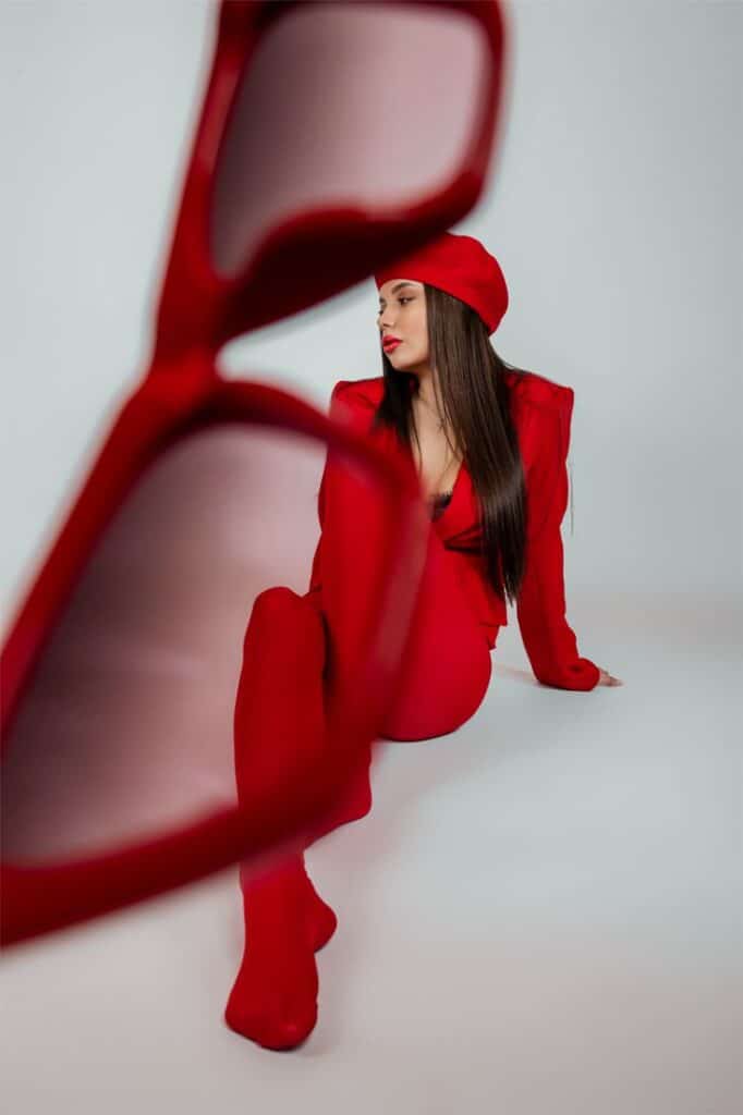 Fashionable woman in red outfit and beret viewed through red sunglasses, posing against a white background.