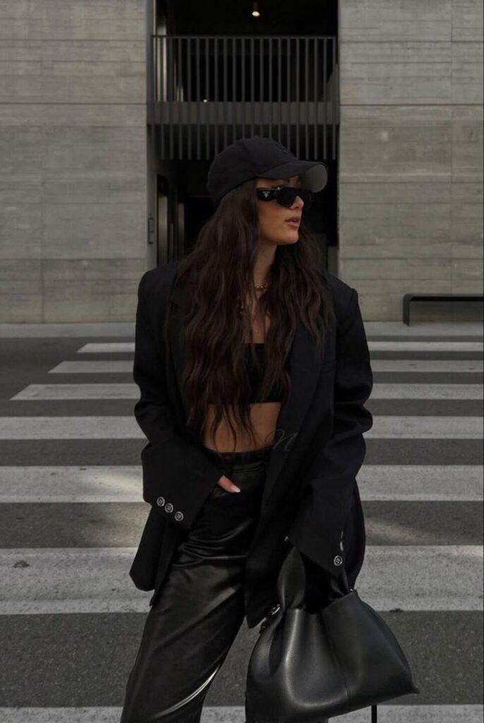 Fashionable woman in black outfit with hat and sunglasses, standing on a city street crosswalk, holding a handbag.