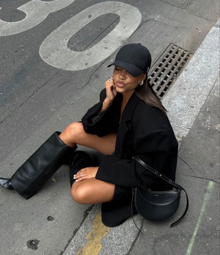 Woman in black outfit and cap sitting on a street curb, stylishly posing with a purse and boots.