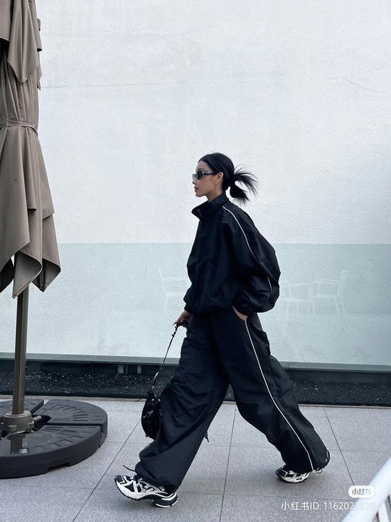 Woman in stylish black outfit and sneakers walking by umbrella on a street, showcasing modern urban fashion.