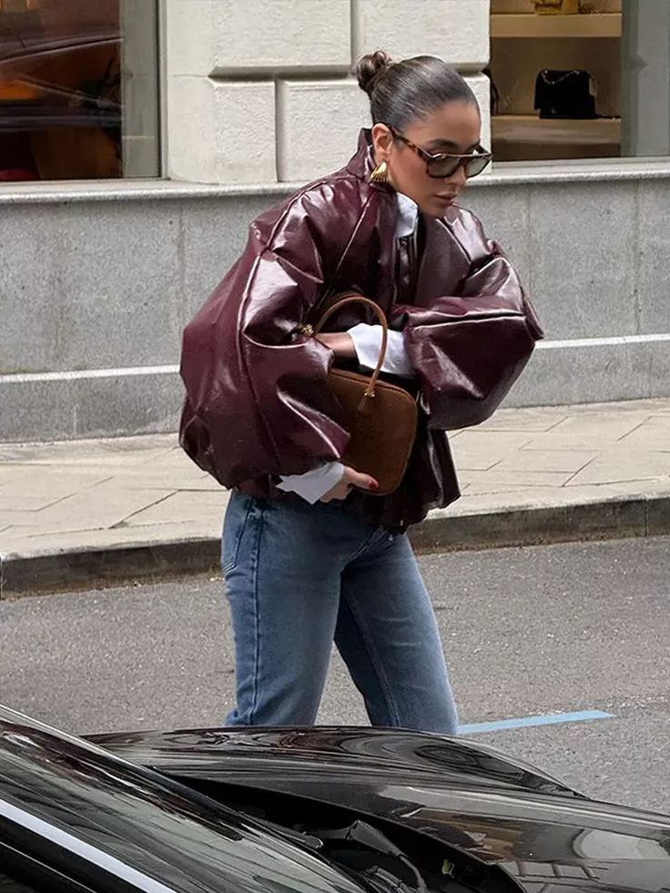 Stylish woman in oversized leather jacket and jeans walking with a brown handbag on a city street.