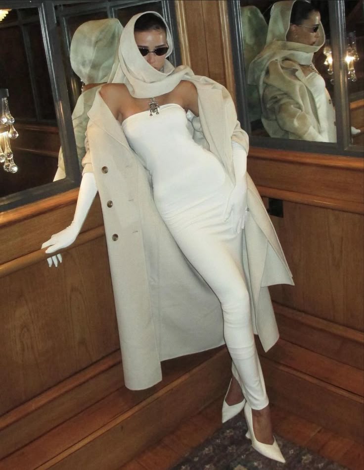 Stylish woman in white outfit and beige coat, wearing sunglasses, poses by mirror in elegant room.