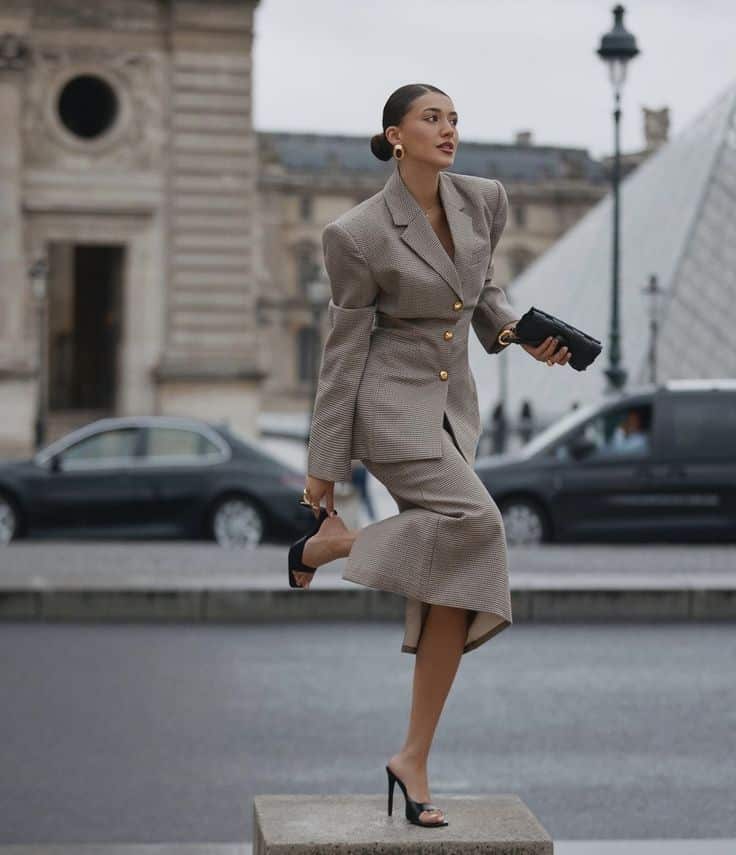 Stylish woman in checkered suit poses confidently near iconic Paris architecture, holding a black clutch.