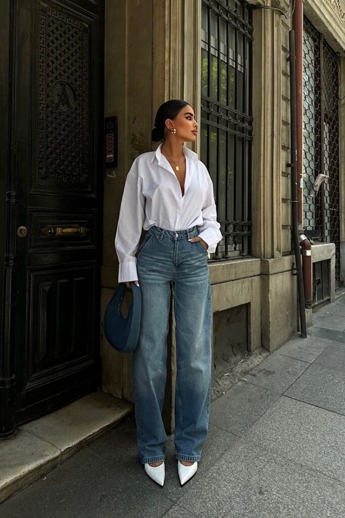 Fashionable woman in white blouse and jeans, holding a blue bag, standing stylishly on a city street.