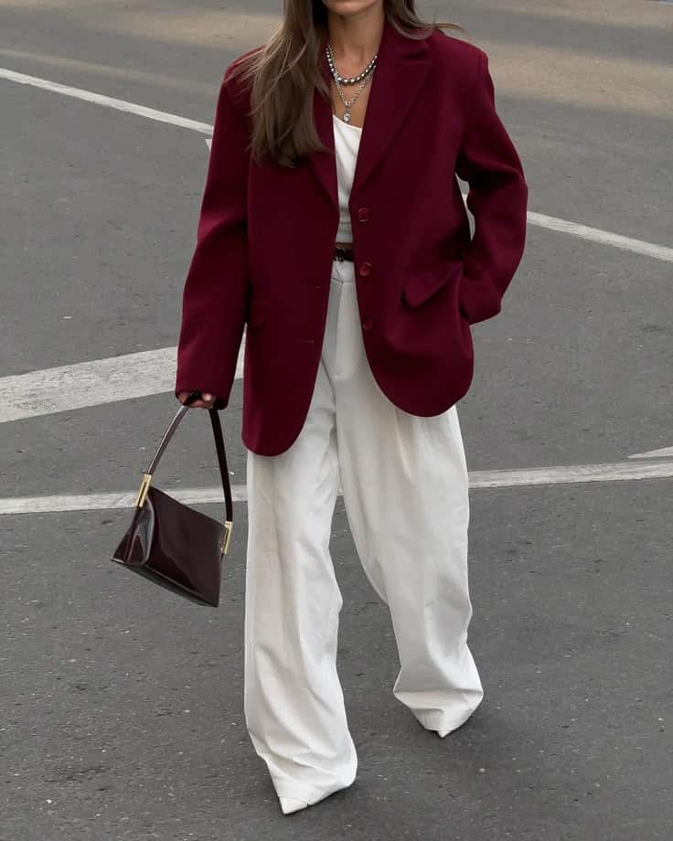 Woman in burgundy blazer and white trousers, holding a matching purse, walking on a city street.