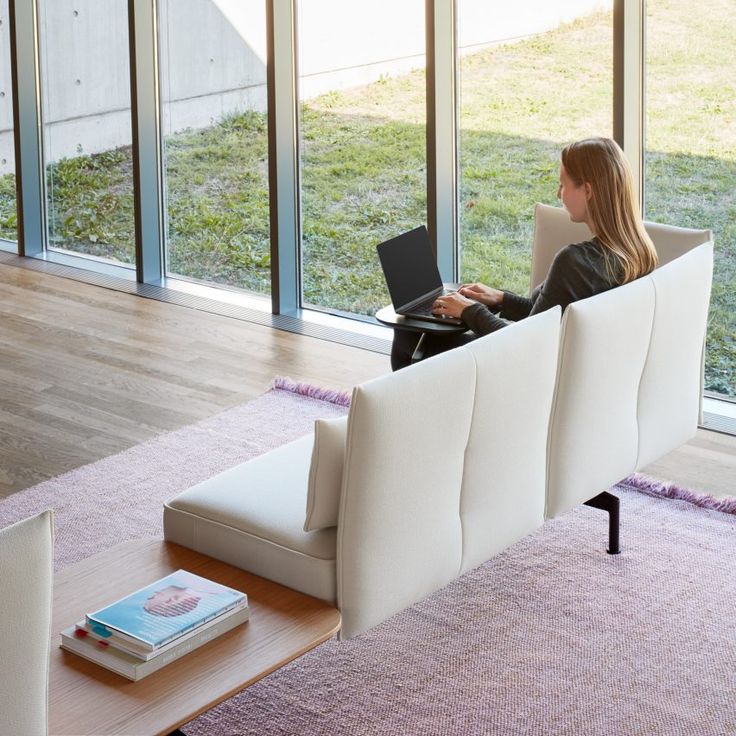 Woman working on a laptop in a bright room with large windows and modern seating, creating a cozy work environment.