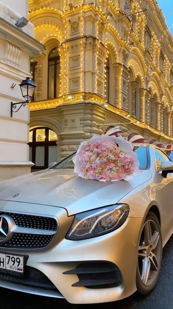 Silver Mercedes parked in front of ornate building with lights, bouquet of pink flowers on hood.