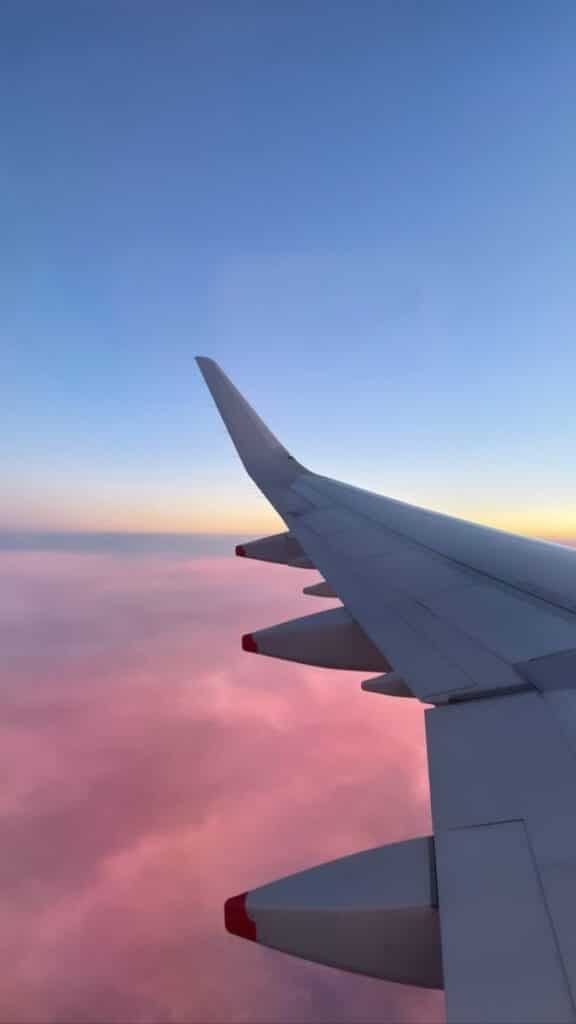 Airplane wing over pink clouds at sunrise, clear blue sky.