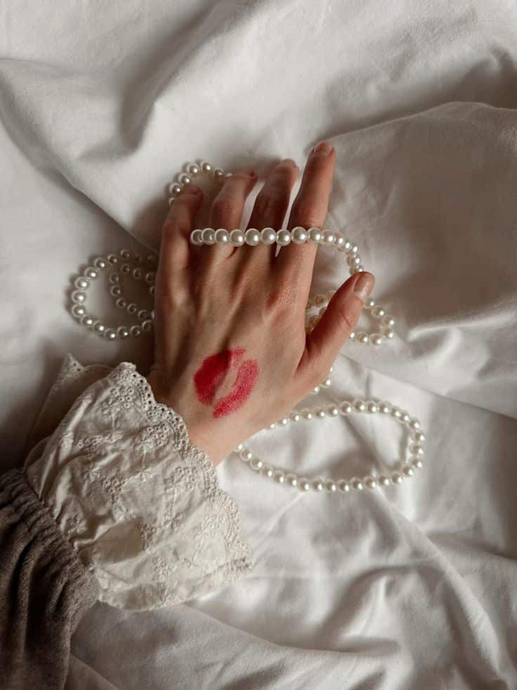 Hand with red lipstick mark holding pearls on white lace fabric.
