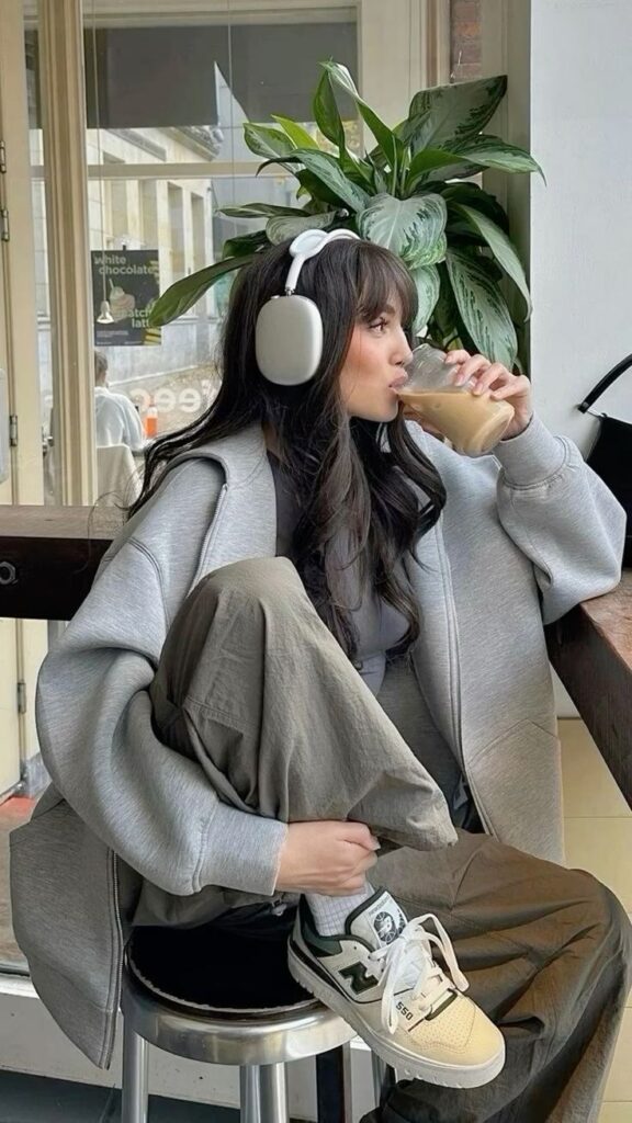 Young woman in cozy attire, sipping iced coffee, wearing headphones, relaxed in a cafe with lush indoor plant.