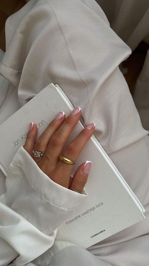 Hand holding a book, wearing rings, with French manicured nails and satin clothing, conveying elegance.