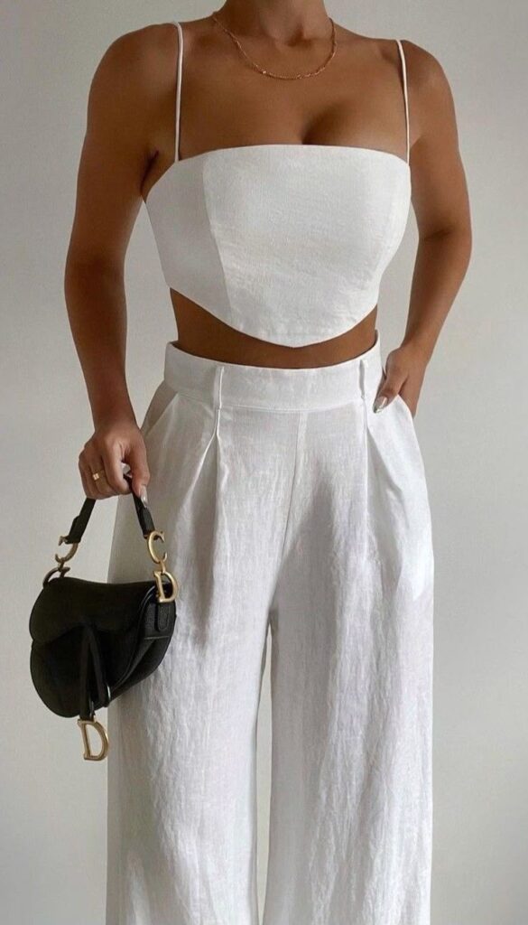Elegant woman in white crop top and pants holding a black designer handbag, showcasing minimalist fashion style.