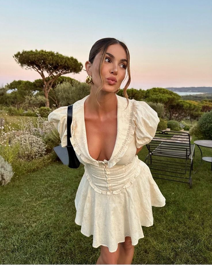 Woman in a stylish white dress poses playfully outdoors with a scenic garden view at sunset.