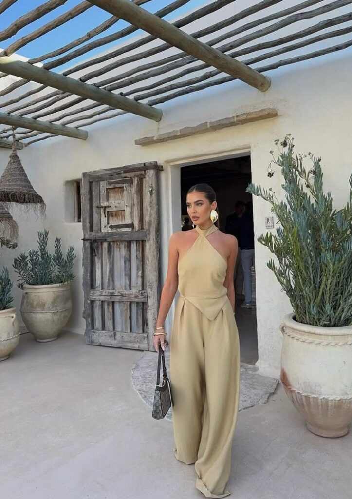 Woman in beige jumpsuit and earrings, exiting rustic building with plants, under wooden pergola. Stylish and elegant.