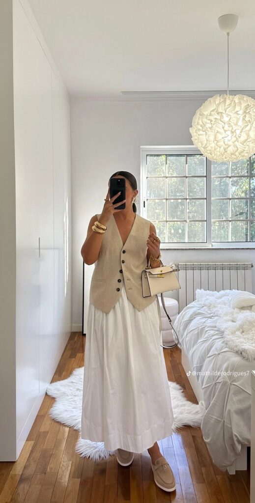 Person in beige vest and white dress takes mirror selfie in bright, cozy bedroom with wood floor and stylish decor.