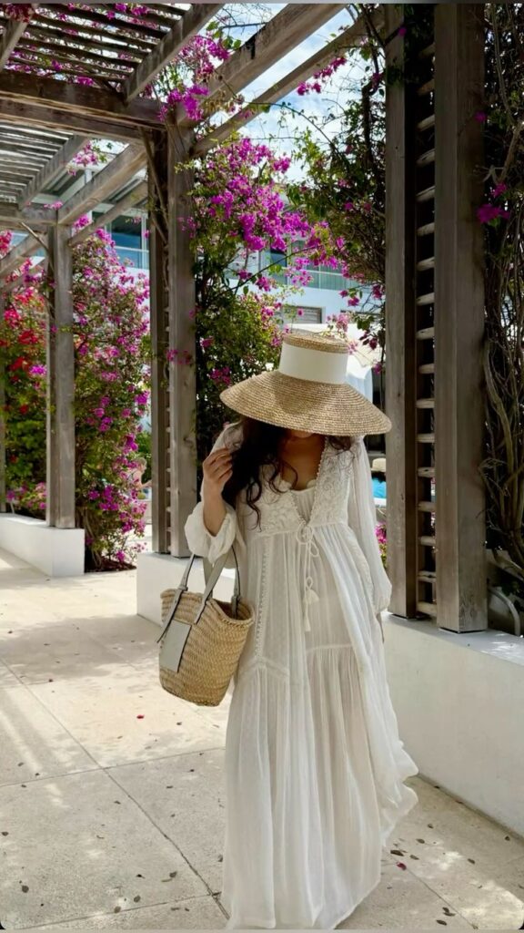 Woman in white dress and straw hat walking under pergola with vibrant pink flowers.