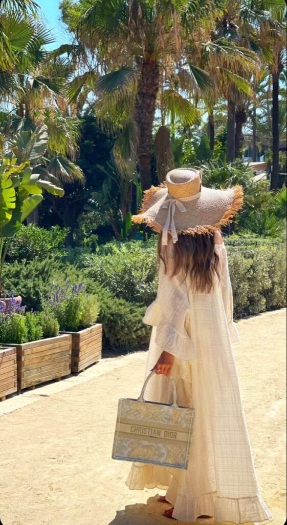 Woman in a flowy dress with a wide-brim hat and Dior bag walking through a tropical garden.