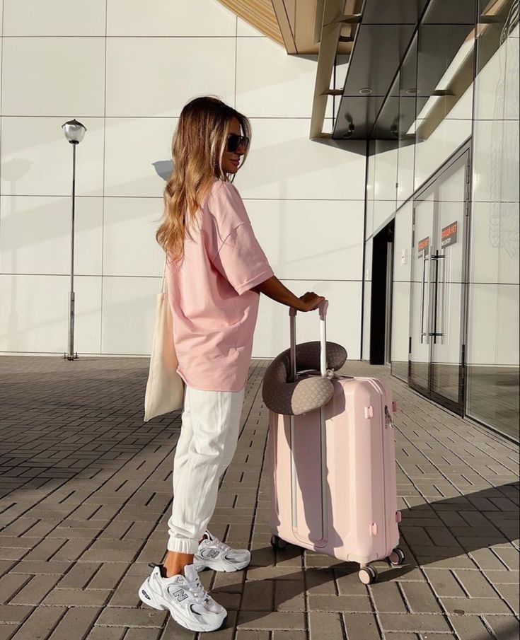 Woman in pink shirt with suitcase and neck pillow outside a modern building, ready for travel.
