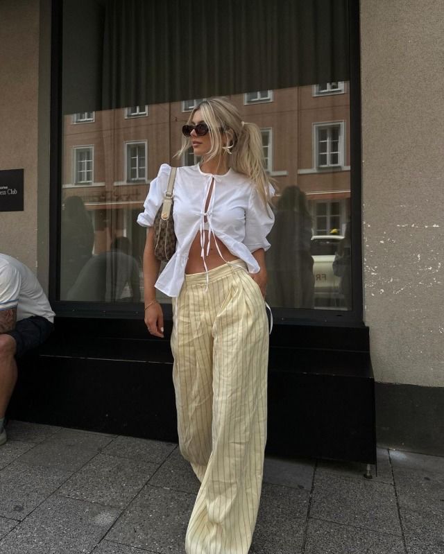 Fashionable woman in a chic outfit with sunglasses, white top, and striped pants standing casually on a city sidewalk.