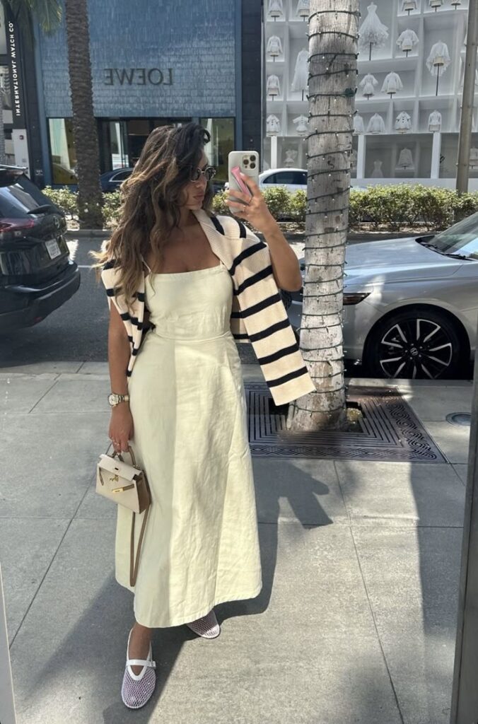 Woman in a chic cream dress and striped sweater, holding a handbag, taking a selfie on a sunny city street. Simple Summer Outfits