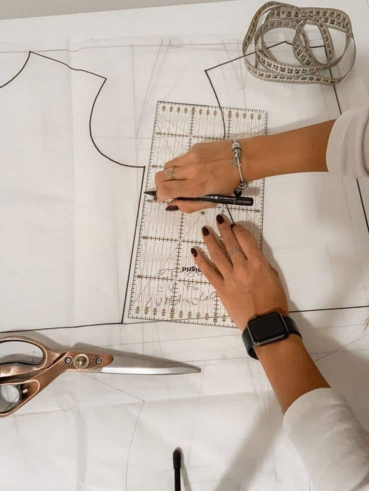 Person measuring and marking sewing pattern with ruler, pen, and scissors on table.