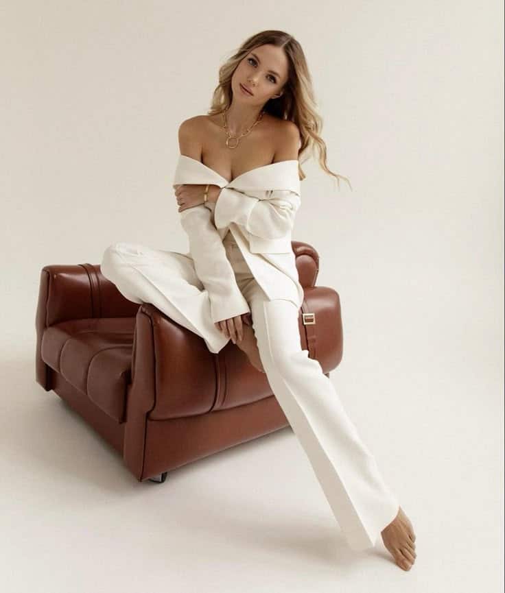 Woman in white suit poses elegantly on brown chair in minimalist setting.