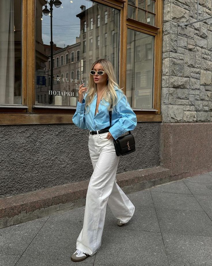 Fashionable woman in blue blouse and white pants walking confidently with sunglasses and a black handbag.