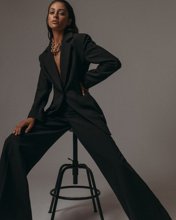 Woman in stylish black suit sitting confidently on a stool, adorned with a chunky gold necklace.