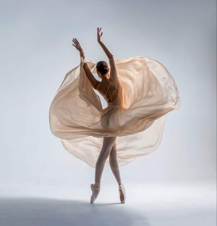 Ballerina in a flowing dress performs an elegant dance pose on pointe against a soft background.