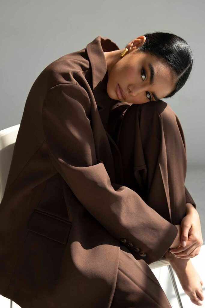 Woman in oversized brown suit sitting on chair, modern fashion portrait with neutral background.